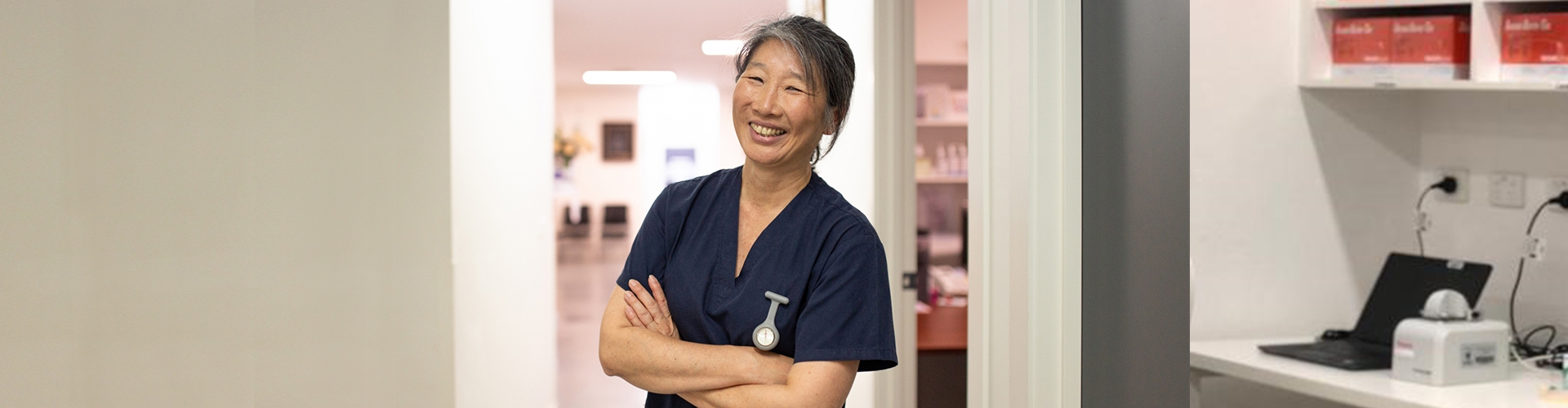 Nurse, wearing navy blue scrubs, smiling and standing in doorway to hospital room with her arms folded across her chest