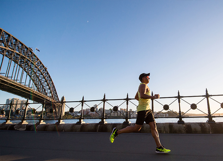 runner wearing a yellow singlet and shoes with black cap and shorts running along Sydney Harbour foreshore with the harbour bridge and city skyline in the background