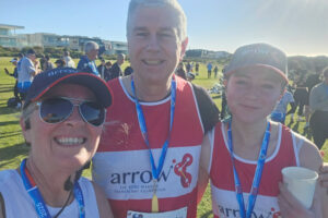 Charlotte Pinney ran the Sutherland to Surf 2025 with her mum and dad and raised over $2000 for Arrow. She is pictured here at the end of the race with her parents. They are all wearing Arrow branded running singlets, and Charlotte and her mum are wearing Arrow branded peak caps.