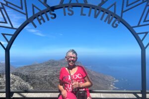 Jenny Nixon, who has learned how to deal with post transplant fatigue, pictured atop a hill in South Africa, standing under a metal arched structure with the words "This is Living" on it. She is looking at the camera, and there is a spectacular vista of land and sea behind her.